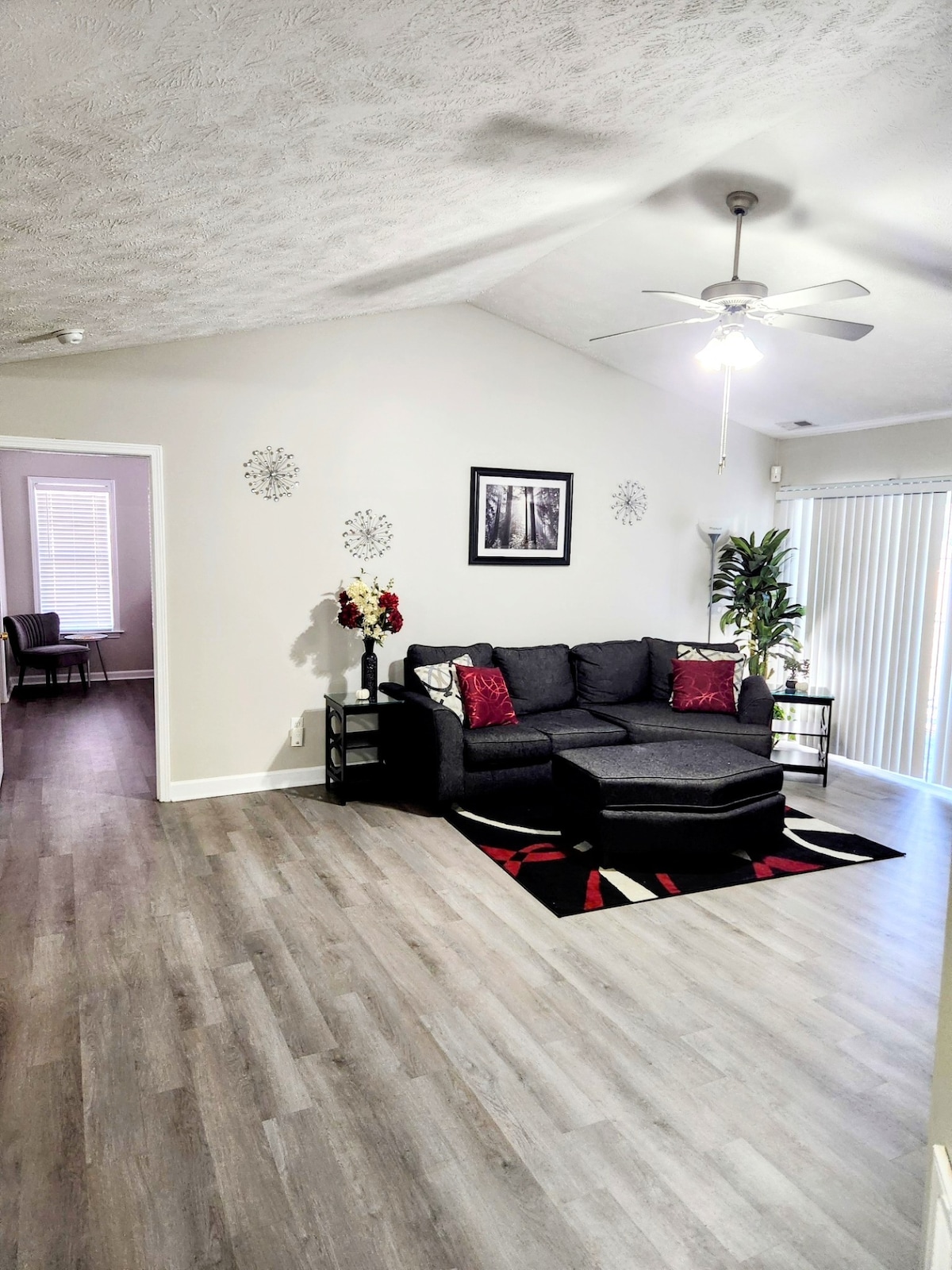 A spacious living area is indicated by high ceilings and soft lighting. A sectional sofa is accented with red pillows, facing a large wall art piece. A decorative rug adds texture to the floor, while a plant adds a touch of greenery. Sliding glass doors provide access to the outside.