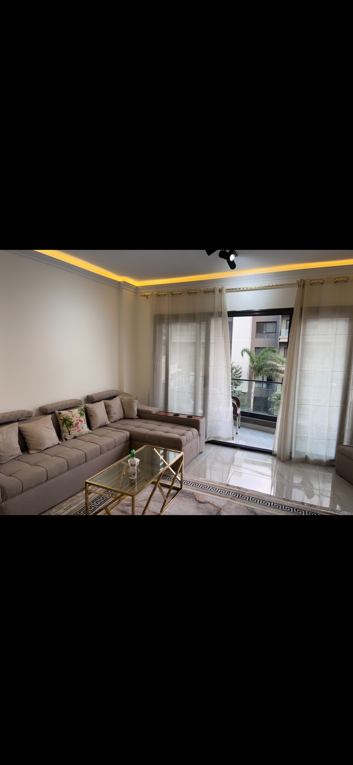 A comfortable living area is highlighted by a spacious sectional sofa, complemented by a glass coffee table. Natural light filters through sheer curtains, illuminating the space. Greenery is visible outside through large glass windows, enhancing the connection to the outdoors.