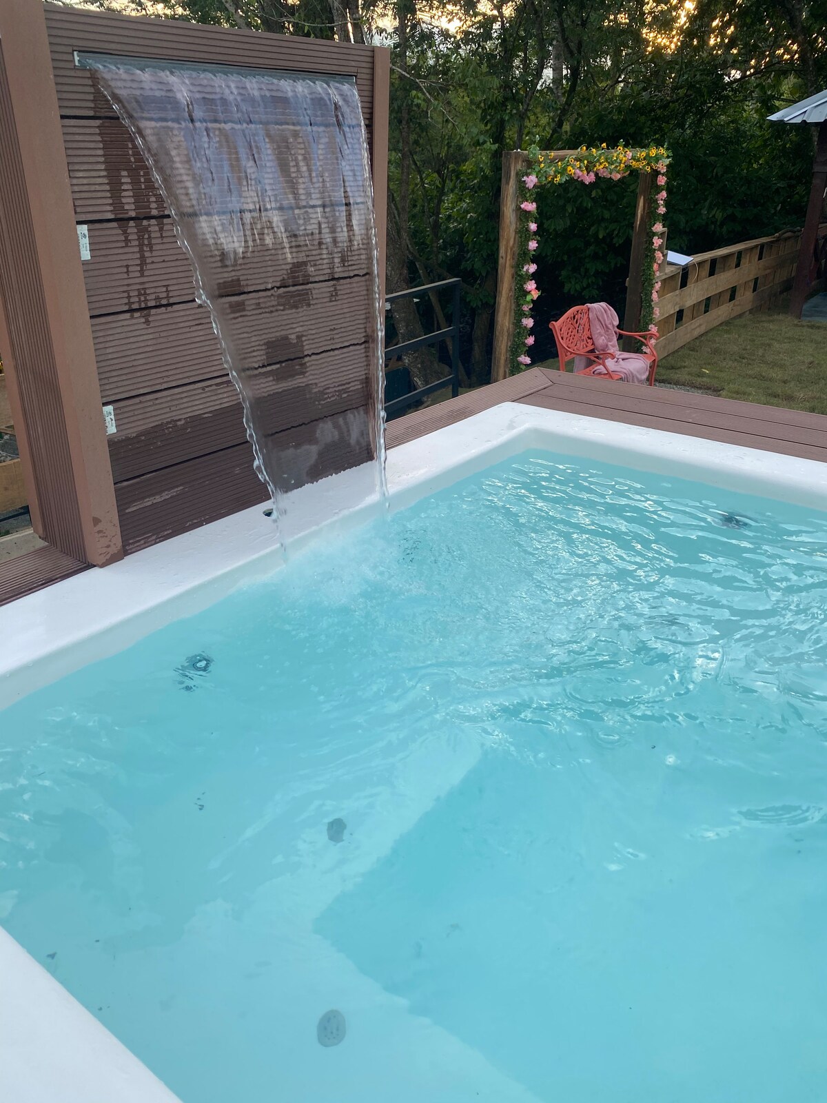 A tranquil pool features clear blue water with a gentle waterfall cascading from a wooden panel. The area is surrounded by greenery, and a lounging chair can be seen nearby, inviting relaxation in the peaceful outdoor setting.