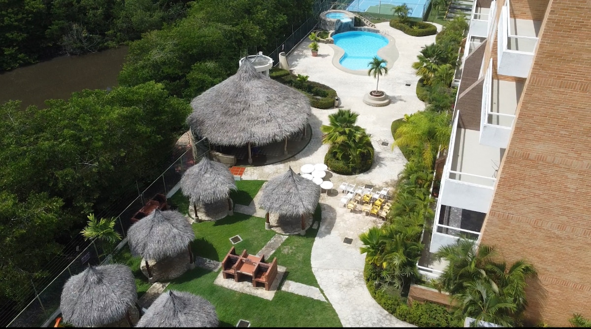 Aerial view of a landscaped outdoor space showcasing a swimming pool surrounded by lush greenery. Thatched-roof shelters and seated areas are arranged on well-manicured lawns, providing shaded spots for relaxation. A barbecue area and palm trees are visible, contributing to a tropical atmosphere.