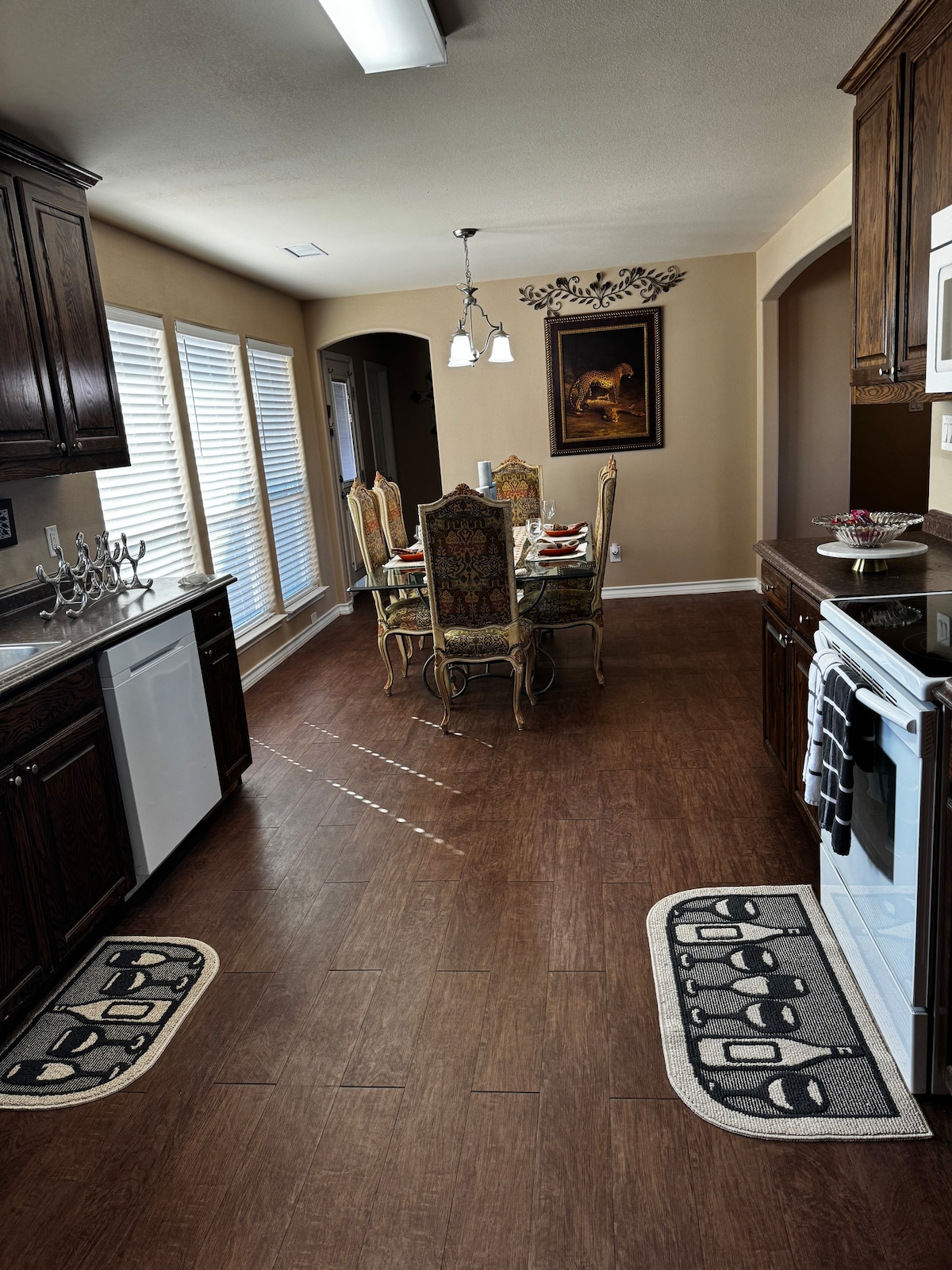 A modern kitchen features dark cabinetry and gleaming countertops. A dining area is set with an elaborate table and upholstered chairs, illuminated by natural light streaming through window treatments. Decorative elements enhance the space, complemented by patterned mats on the floor.