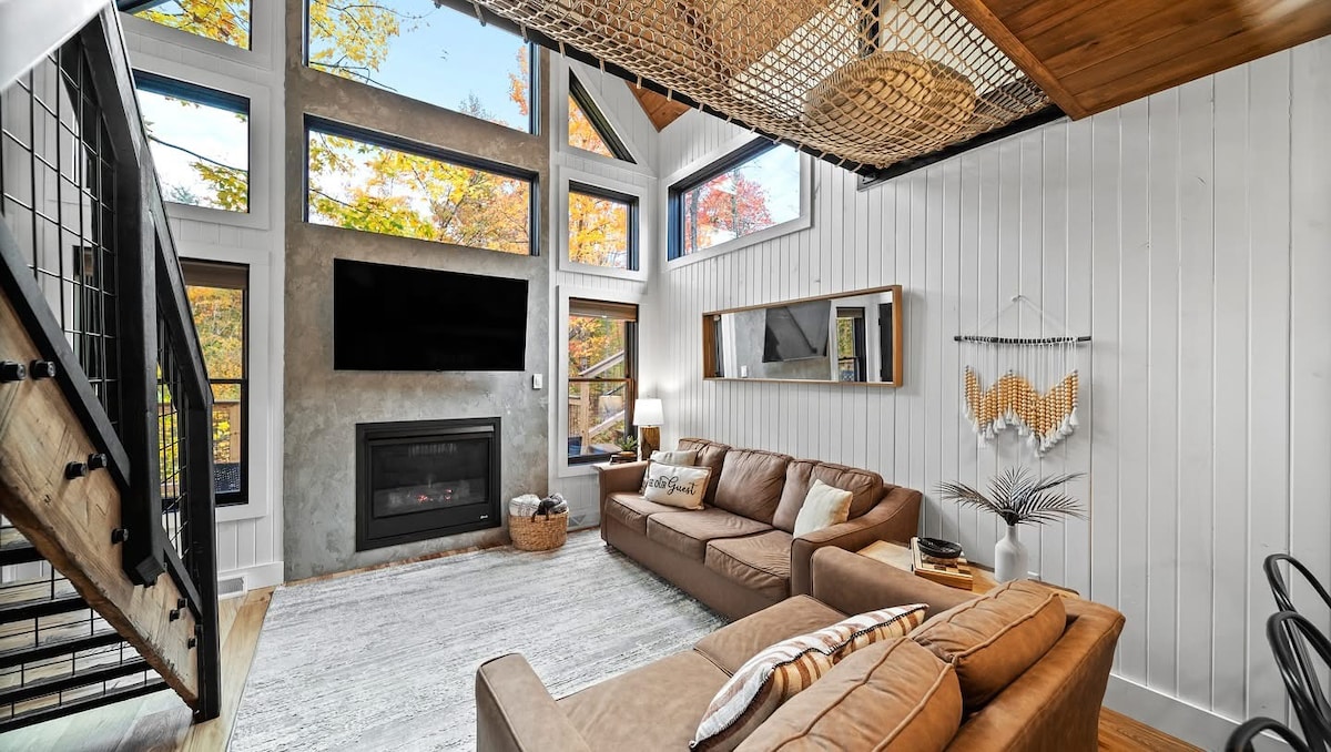 The Overstory — Bright living room with oversized forest-view windows and a gas fireplace.