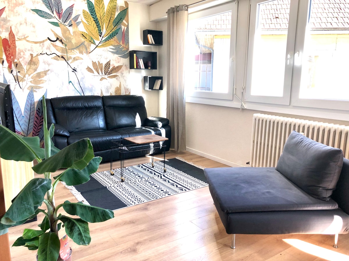 The living area features a dark leather sofa paired with a contemporary lounge chair, situated on a patterned rug. Large windows illuminate the space, showcasing wall art adorned with botanical motifs. A small coffee table and a bookshelf enhance the room's functionality.