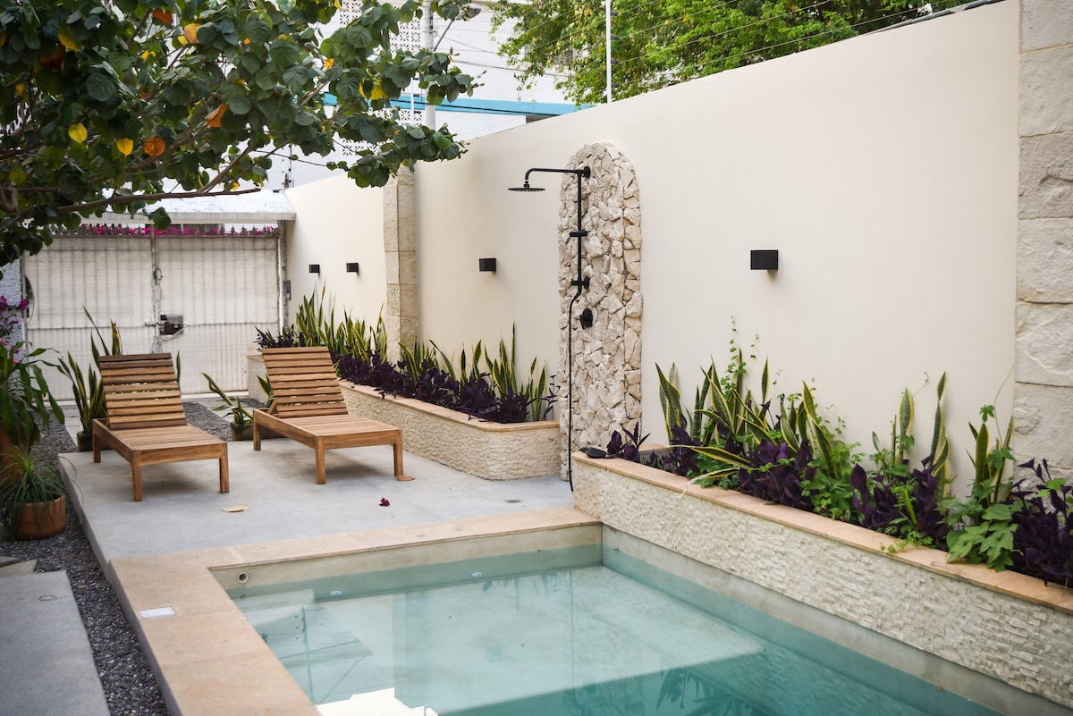 An inviting garden area features a small pool surrounded by a laid-stone pathway. Two wooden lounge chairs are positioned nearby, complemented by lush green plants along the walls. Soft lighting is provided by wall-mounted fixtures, enhancing the serene environment.