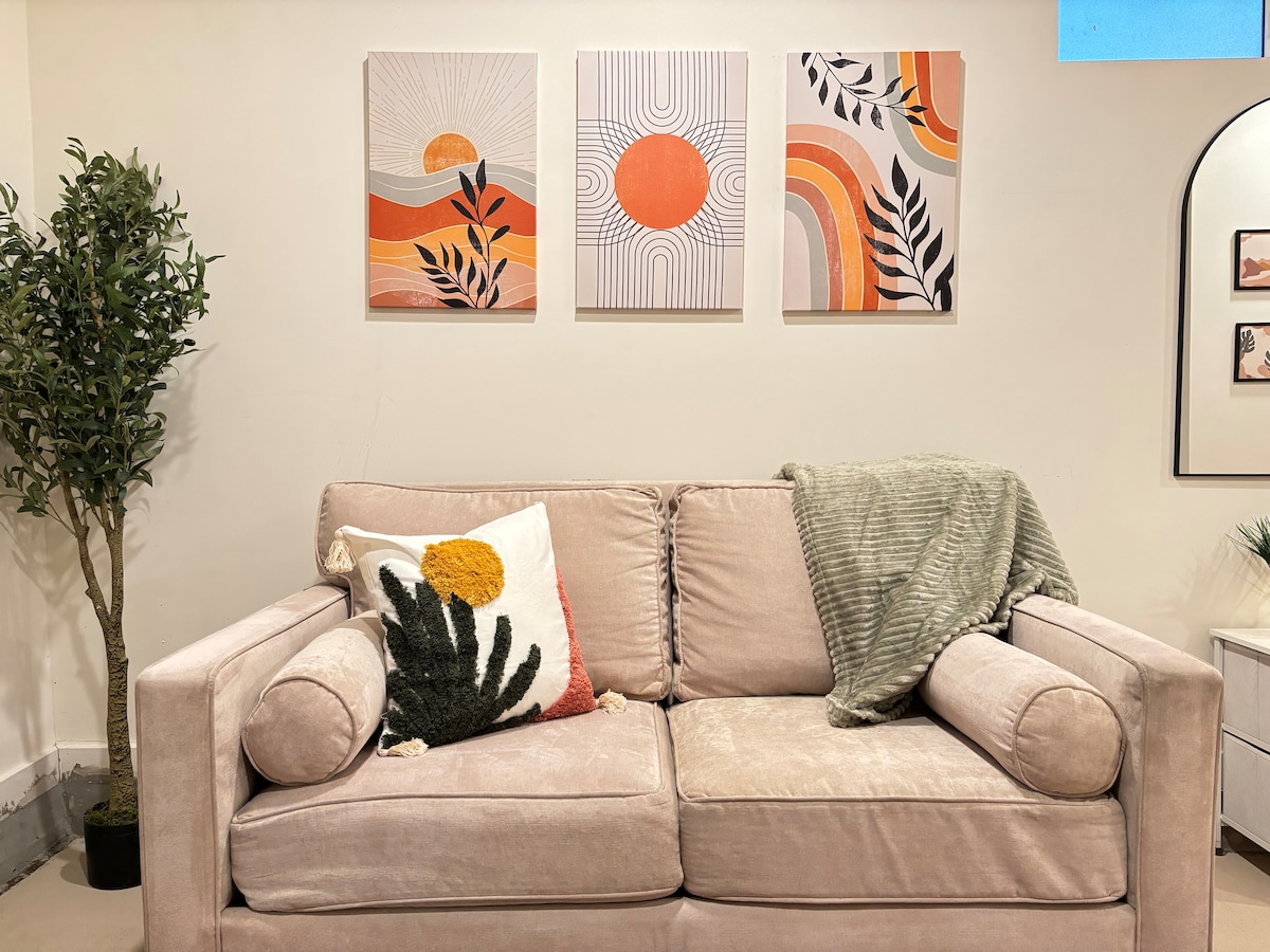 A soft beige couch features decorative pillows and a cozy throw blanket. Above the couch, three abstract art pieces display warm colors and organic shapes. A green plant is positioned beside the couch, adding a touch of greenery to the space.