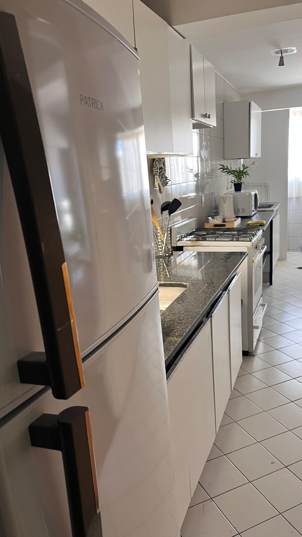 A modern kitchen is presented with sleek white cabinetry and a dark granite countertop. Stainless steel appliances, including a refrigerator and oven, are visible. Neutral tile flooring runs throughout, with natural light filtering in from a nearby window. A small plant adds a touch of greenery.