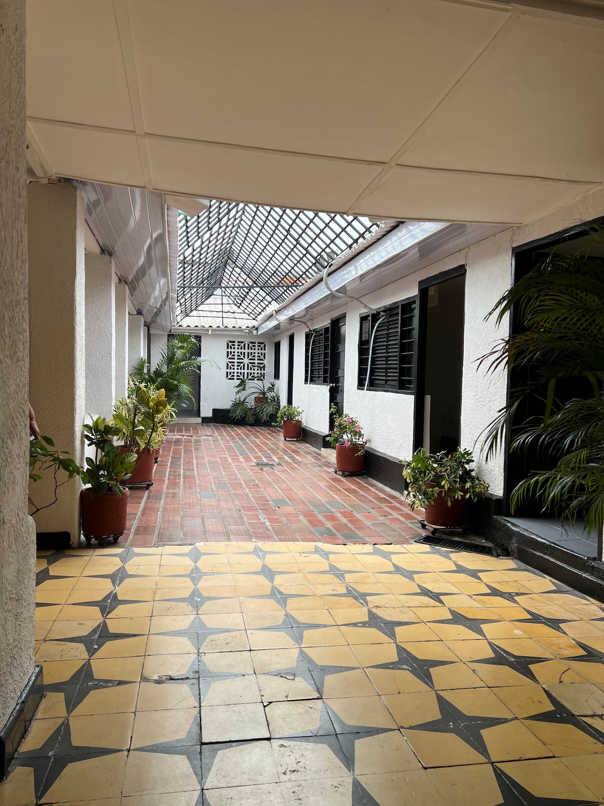 A spacious corridor is lined with potted plants and features a tiled floor with a geometric pattern. Natural light filters through the glass roof, illuminating the area. Doorways lead to adjoining rooms, providing a glimpse of the tranquil environment.