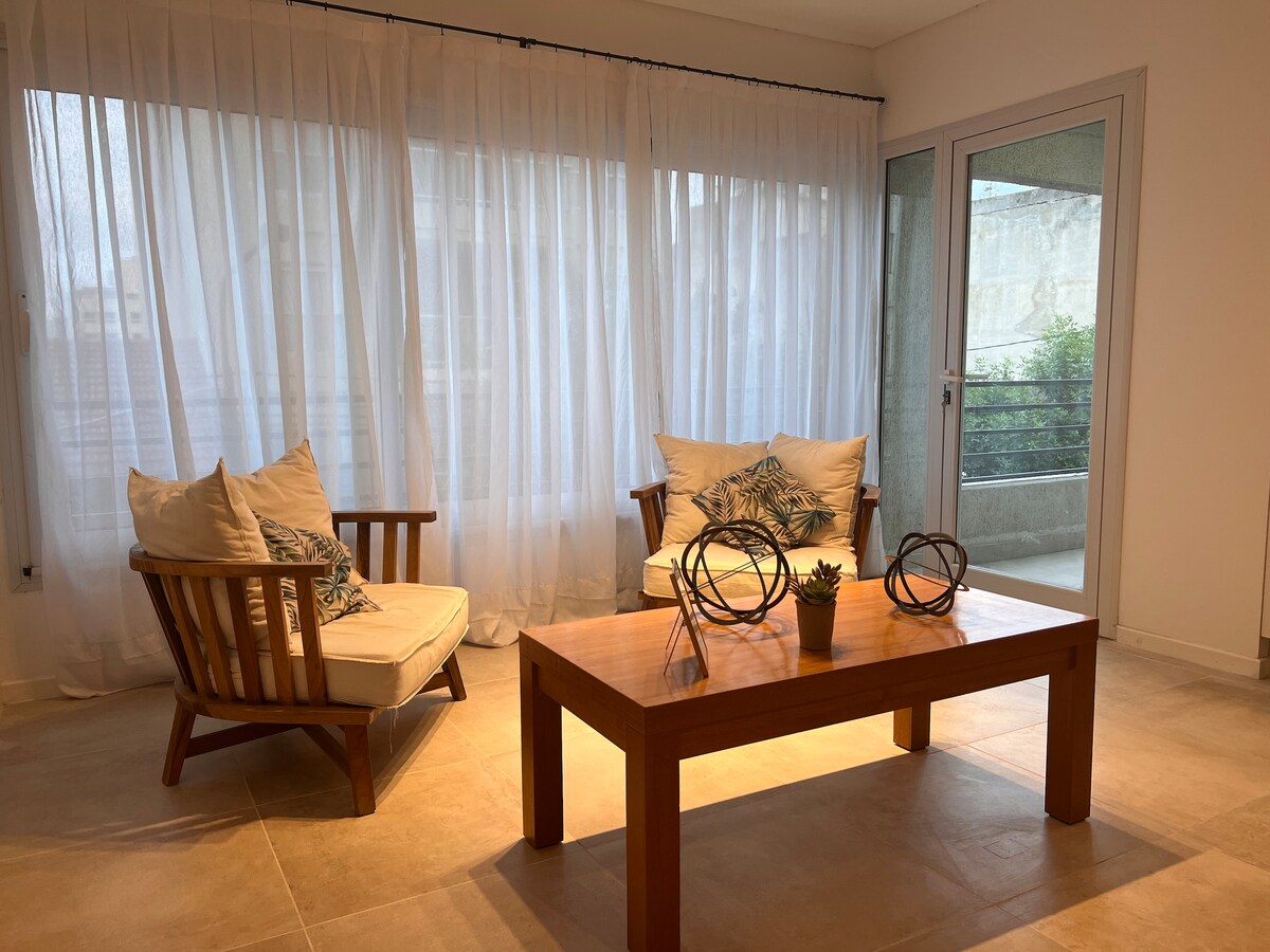 A bright and spacious living area features two wooden chairs with soft cushions, positioned beside a wooden coffee table. Natural light filters through sheer curtains covering large windows, while a small plant and decorative items are arranged on the table.