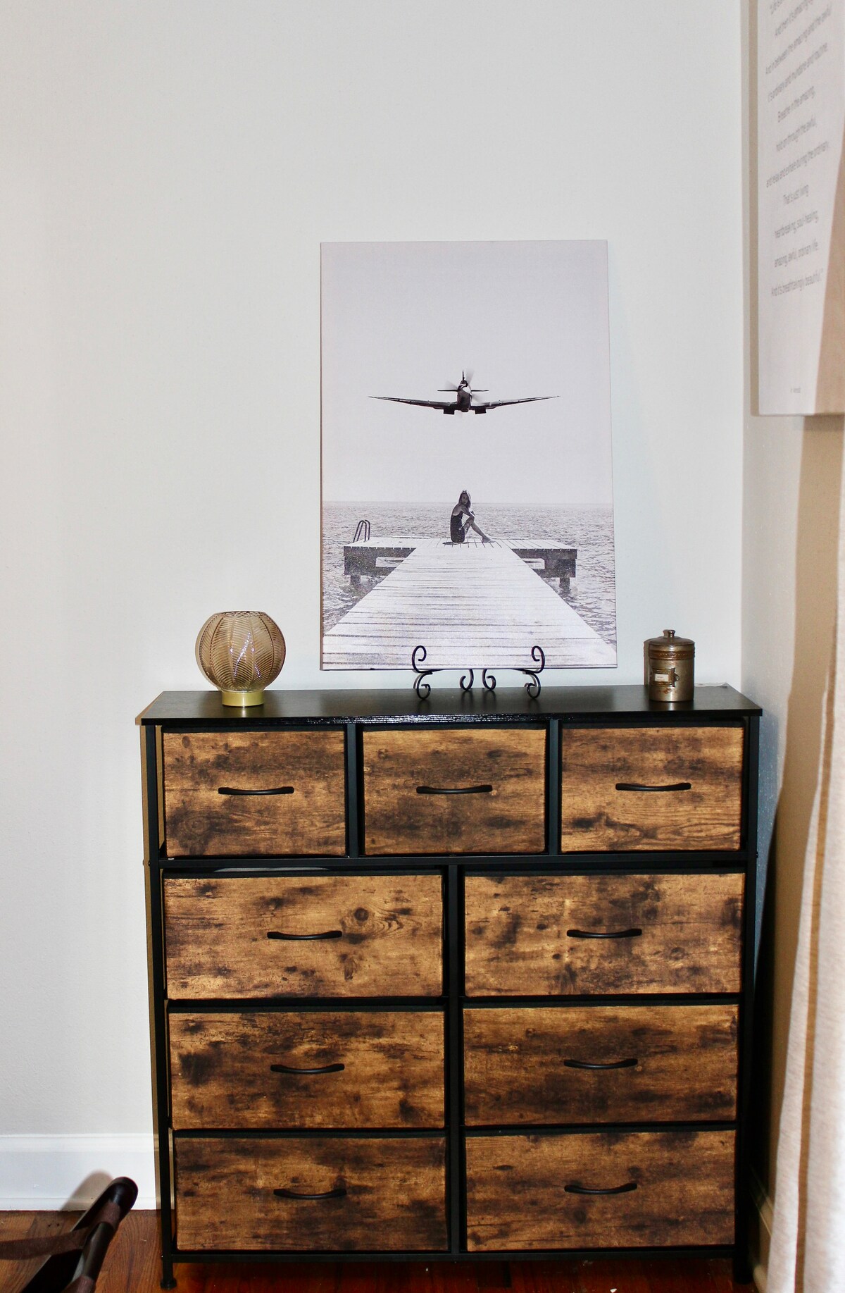 A sturdy chest of drawers displays a blend of natural wood and black metal, providing ample storage. Above it, a monochrome artwork features a serene scene of a person on a dock with an airplane overhead. Subtle decorative items are placed on the chest.
