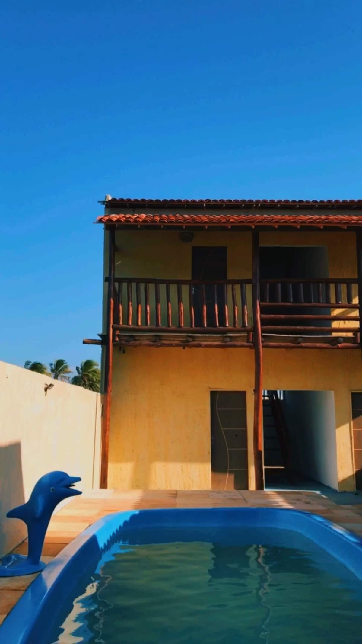 A two-story building is displayed, featuring a traditional design with a sloped roof and wooden balconies. A bright blue dolphin sculpture is positioned near a pool, which reflects the clear blue sky overhead. The surrounding area is spacious and inviting.