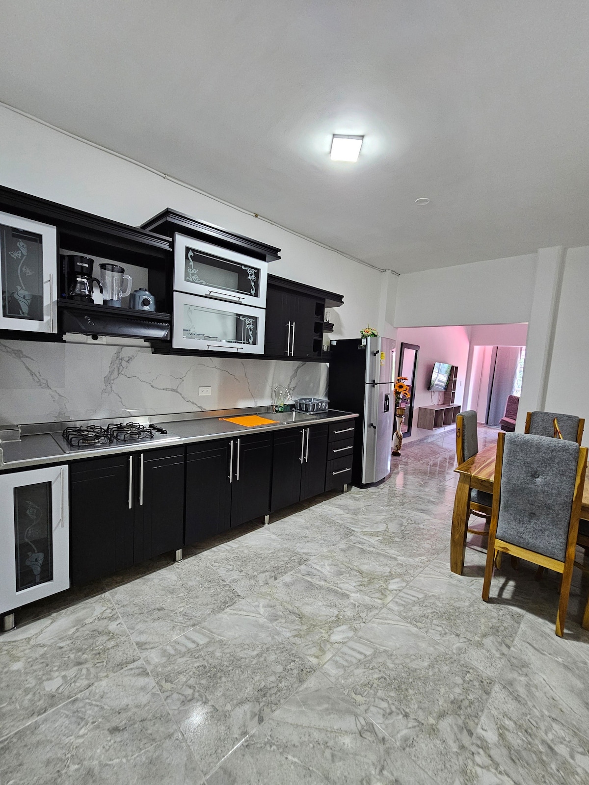A modern kitchen is presented with sleek black cabinetry and granite countertops. Stainless steel appliances, including a refrigerator, are visible, along with a six-person dining table made of light wood. The space is illuminated by overhead lighting, and the floor features polished stone tiles.