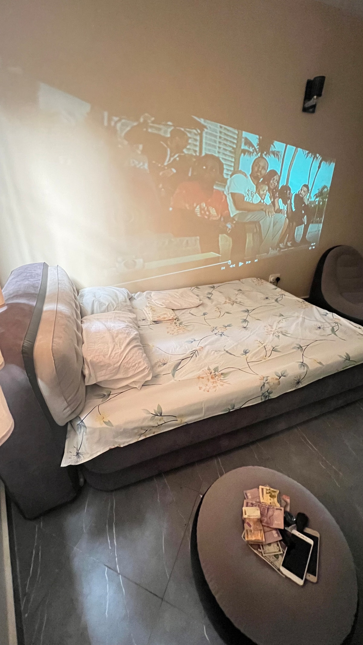 A modern sleeping area features a spacious bed with a floral-patterned blanket. A video projector displays an image on the wall, adding to the ambiance. Nearby, a round coffee table holds a few items, while soft seating completes the cozy atmosphere.