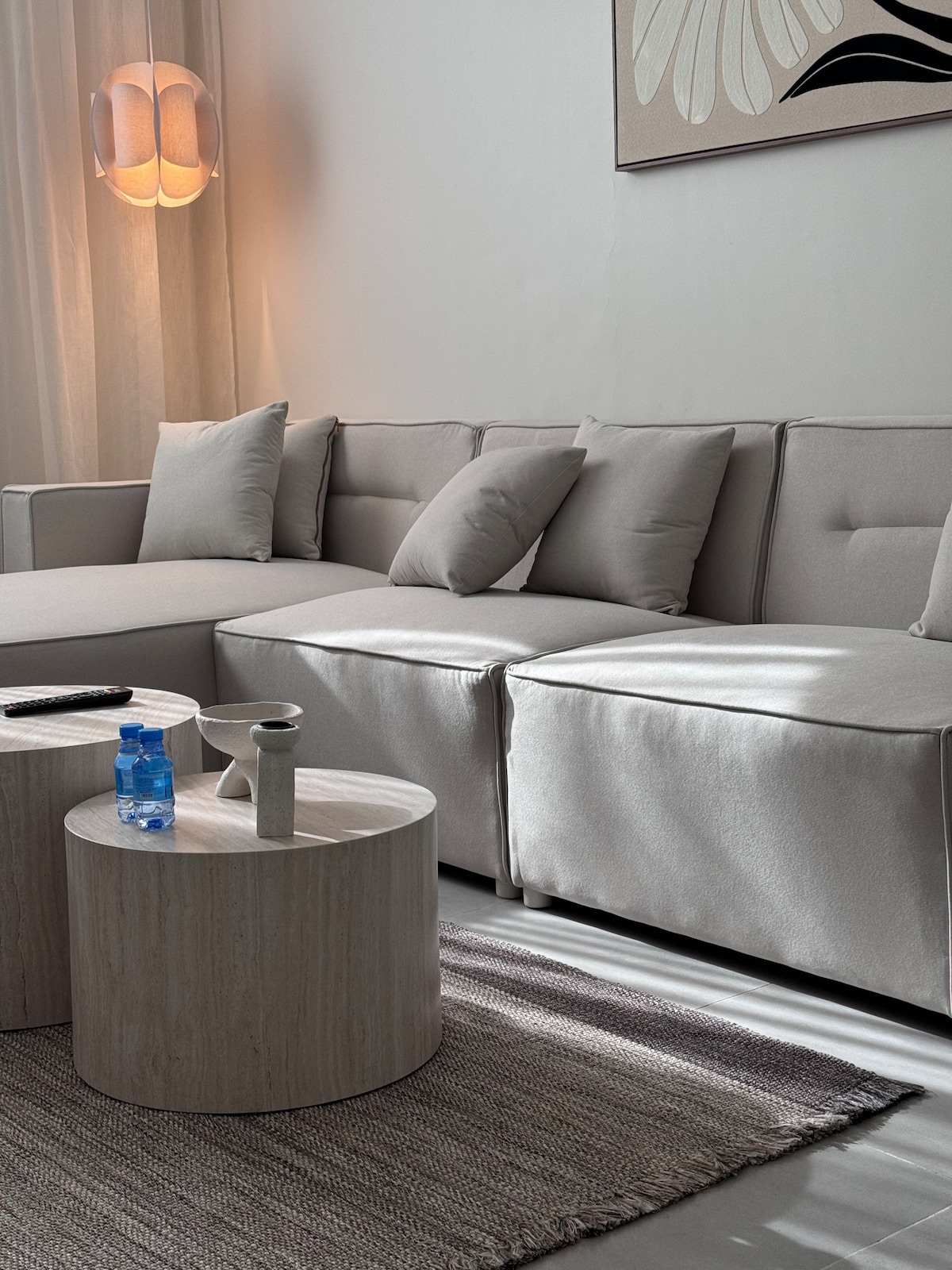 A modern lounge area is presented, featuring a light-colored sectional sofa adorned with plush cushions. Two circular tables in natural tones sit adjacent, holding a water bottle and a small decorative item. Soft light filters in, creating a comfortable and inviting space.