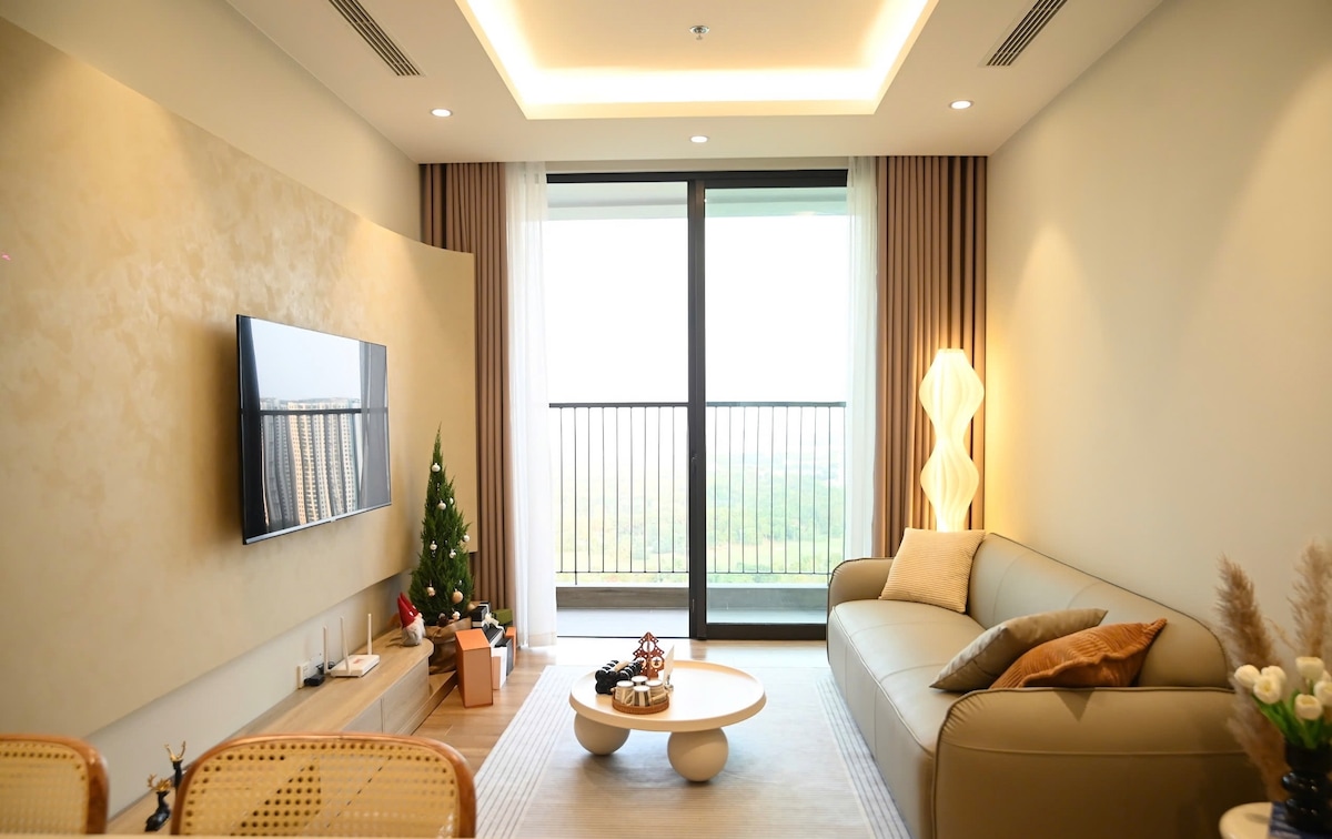 A modern living area features a soft beige sofa and a round coffee table. Natural light streams through a large window, and a small decorative Christmas tree adds a festive touch. The wall is equipped with a flat-screen television, complementing the minimalistic decor.