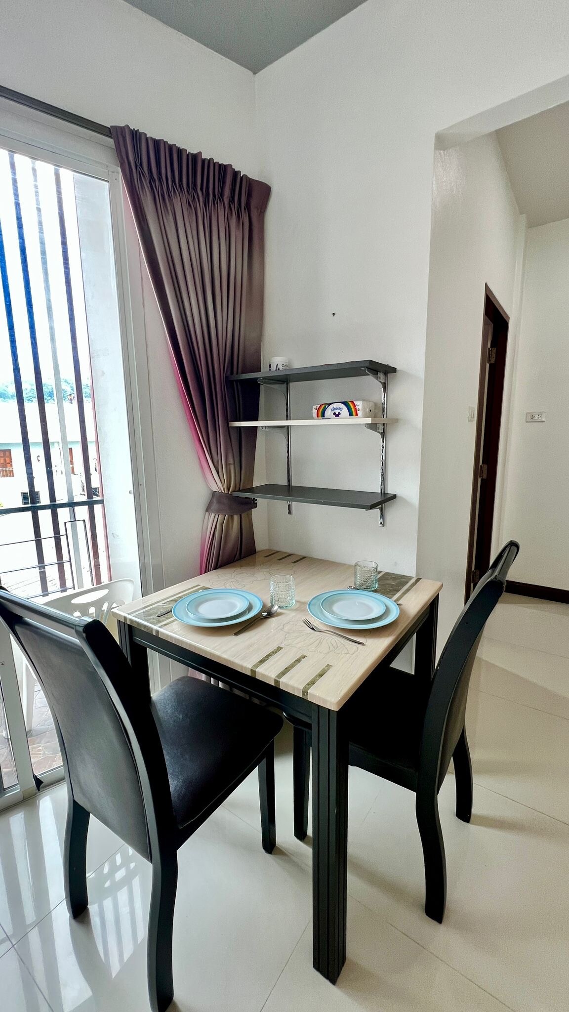 A small dining area is presented with a rectangular table set for two, featuring light-colored plates and glassware. Black chairs accompany the table, and a shelf with decorative items is mounted on the wall. Natural light enters through large glass doors with vertical blinds.