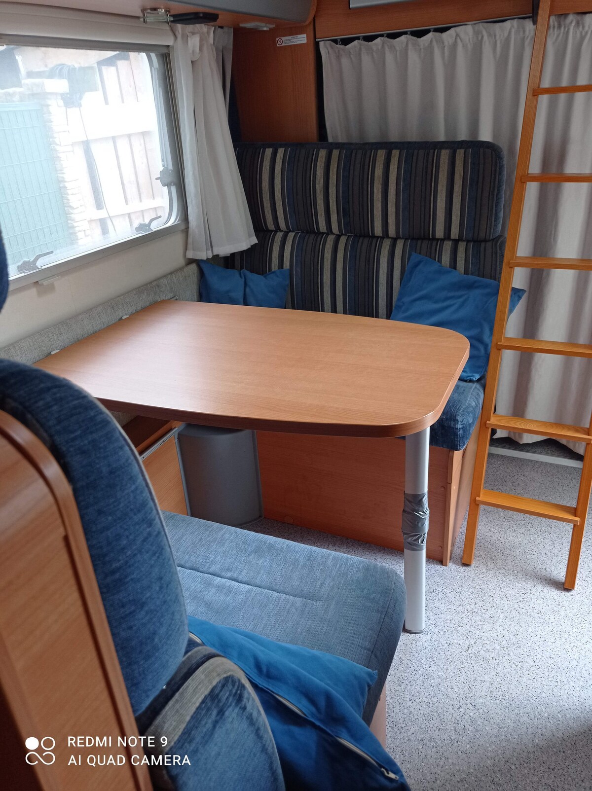 A compact seating area features a light wooden table accompanied by cushioned chairs in patterned fabric. A small ladder is visible near the seating, providing access to an upper sleeping space. Natural light filters in through the window, illuminating the soft blue and beige tones.