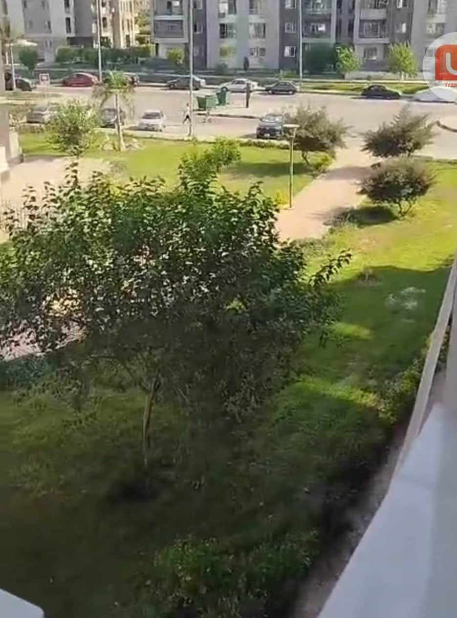 A peaceful garden area is visible, featuring neatly trimmed grass and several small trees. The walkway is surrounded by greenery, providing a serene view. In the background, nearby buildings and a road can be seen, creating a sense of proximity to urban amenities.