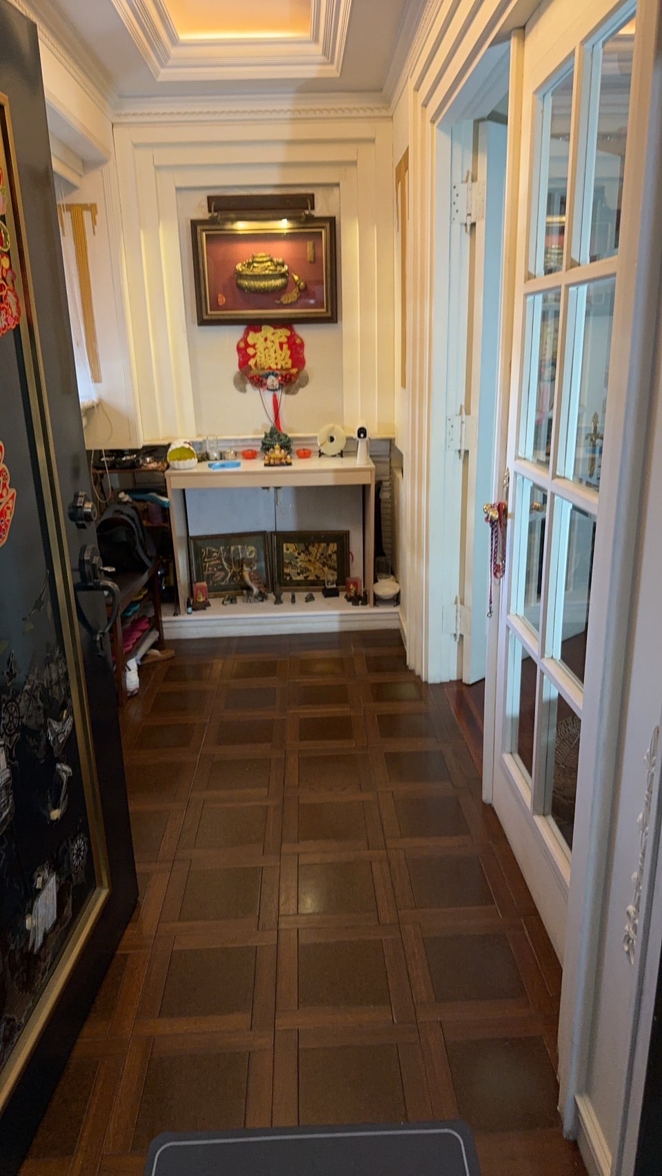 A welcoming entryway features a polished wooden floor and bright white walls. A decorative table with cultural artifacts and a floral arrangement is positioned against the back wall. Glass-paneled doors provide access to another room, enhancing the open feel of the space.