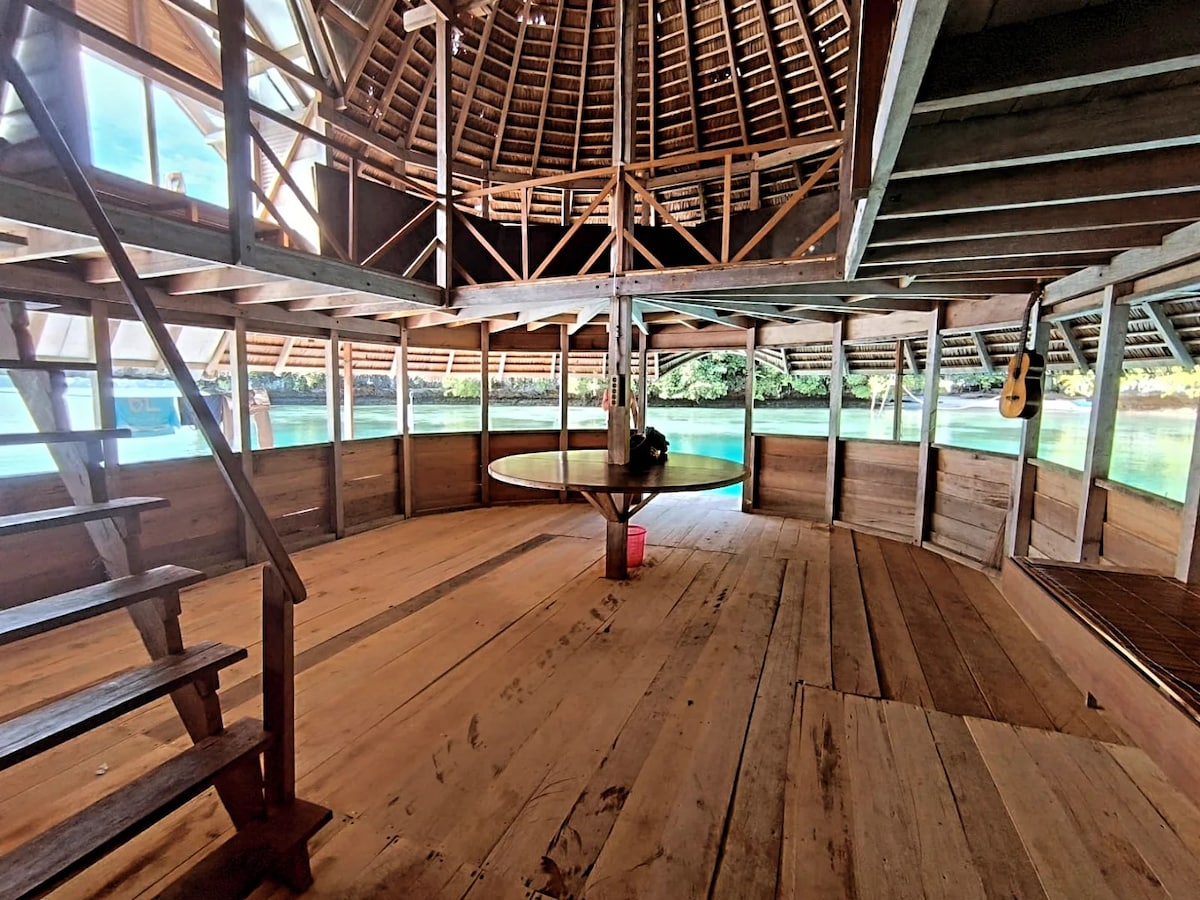 An open and airy interior features a circular wooden design with a high thatched roof. Light filters through large windows, illuminating the wooden floors. A central dining table is positioned in the middle, surrounded by wooden beams and a staircase leading to another level.