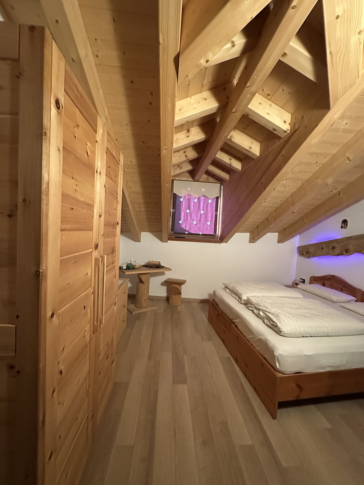 A cozy bedroom features wooden beams and flooring, contributing to a warm ambiance. A spacious bed is positioned under a slanted ceiling, and a small table with two wooden stools is nearby. A window allows natural light to enter, complemented by soft purple curtains.