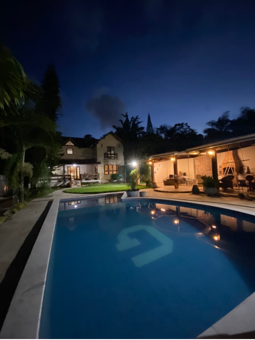 A serene evening scene features a well-lit swimming pool surrounded by lush greenery. The pool reflects the soft glow of ambient lights, while a welcoming outdoor seating area is visible nearby. An elegant home structure stands in the background under a twilight sky.