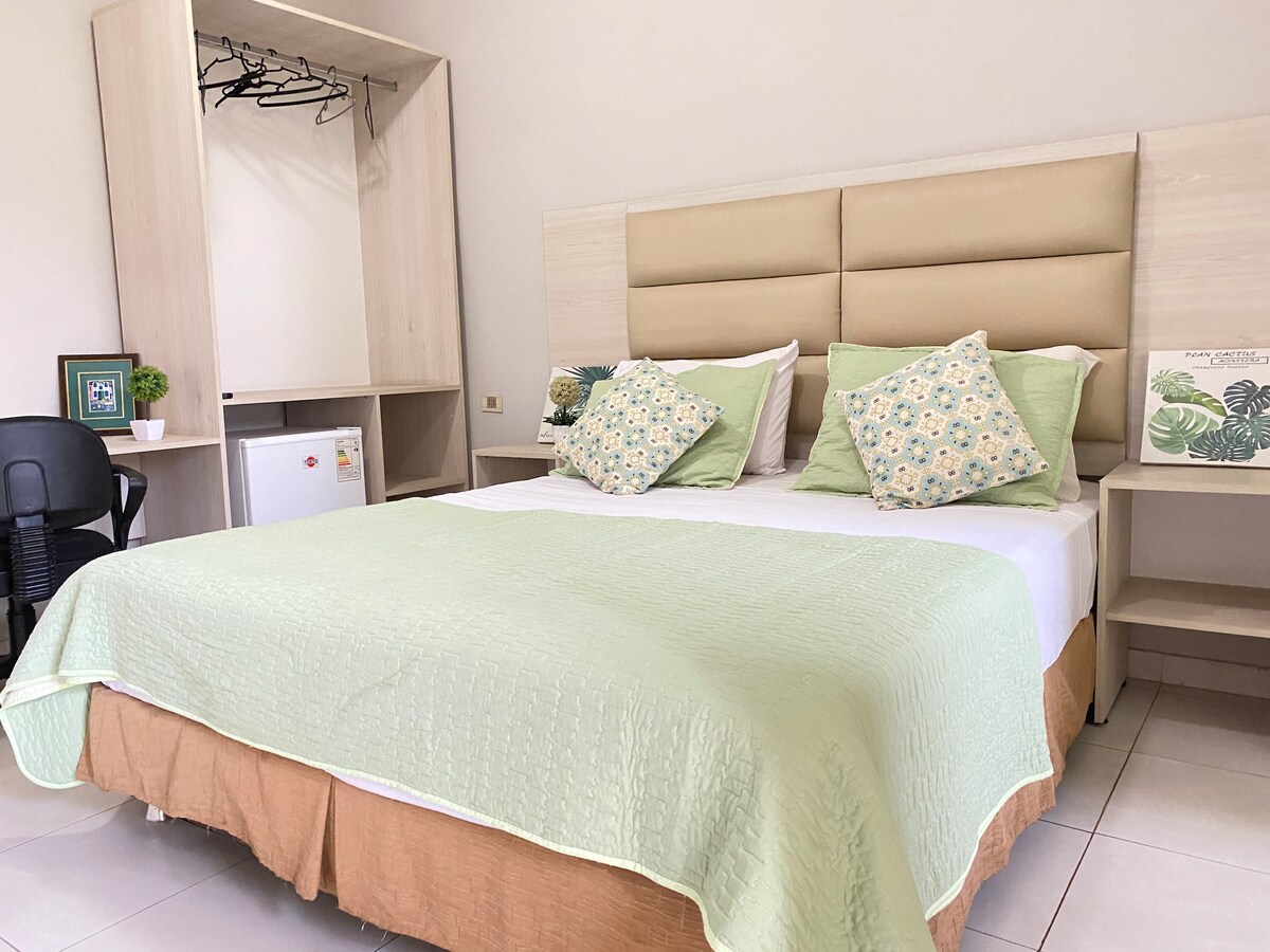 A neatly arranged bedroom features a spacious bed covered with a soft green quilt. Decorative pillows add color to the bed. A minimalist wardrobe and desk accompany a small fridge, providing convenience for guests.