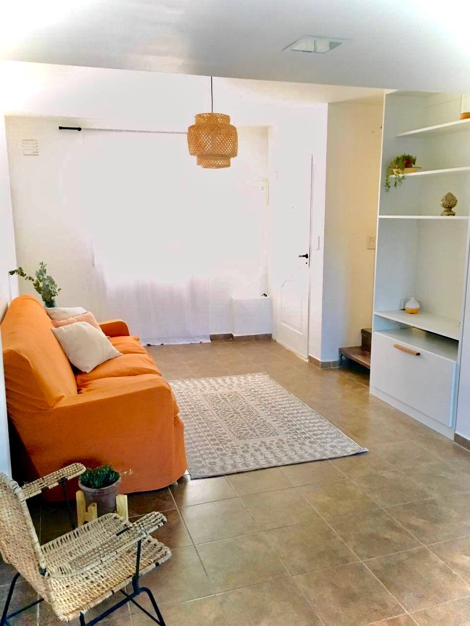 A living area is highlighted by an orange sofa, accompanied by a small chair and a decorative rug. Natural light floods in through a window with sheer curtains, creating a bright and airy space. Shelving units display plants and decor, adding character to the room.