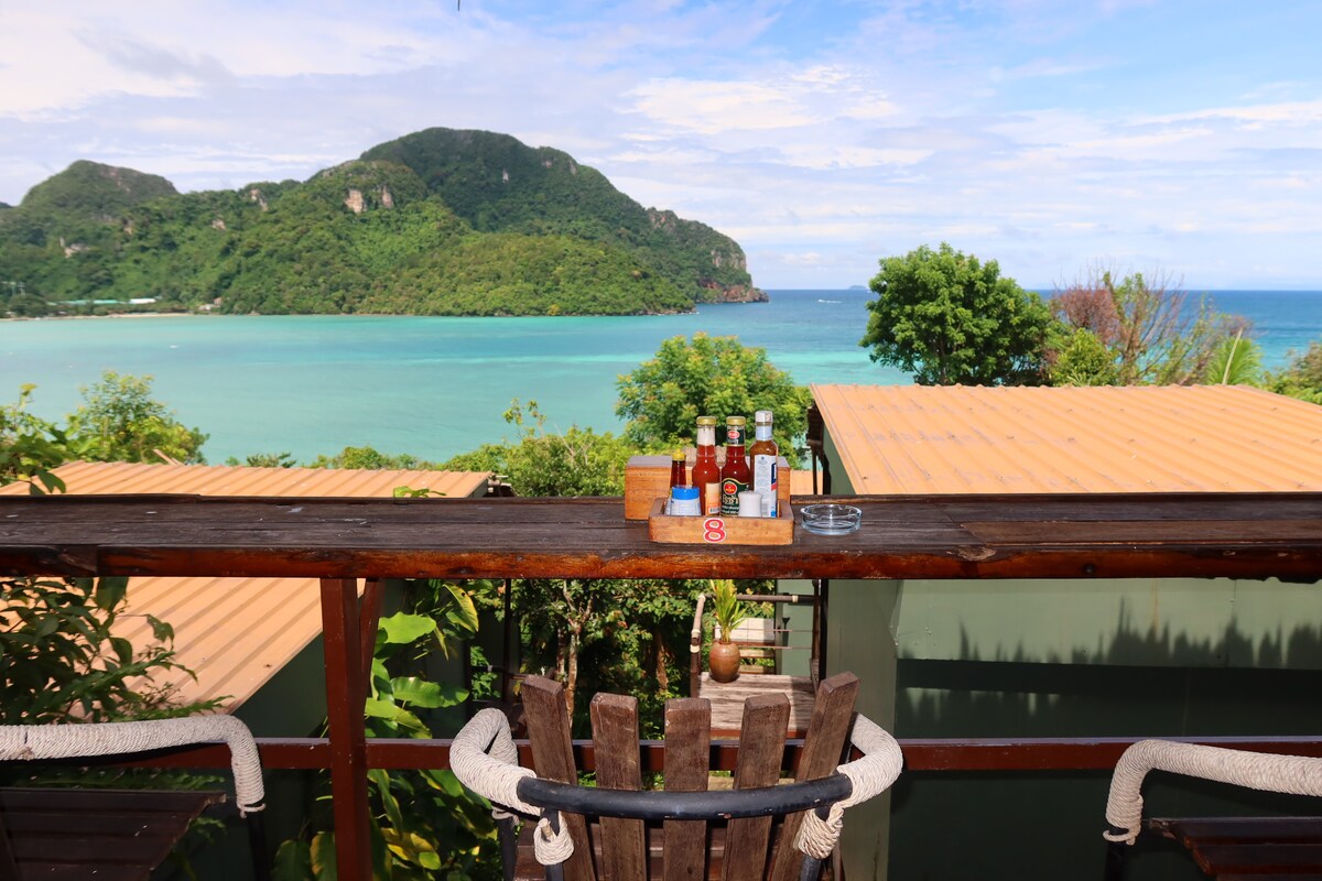 A wooden balcony overlooks a stunning ocean view, framed by lush greenery and distant hills. A wooden table holds a selection of small bottles, with a cigarette ashtray nearby. Bright sunlight enhances the serene setting against the clear blue water.
