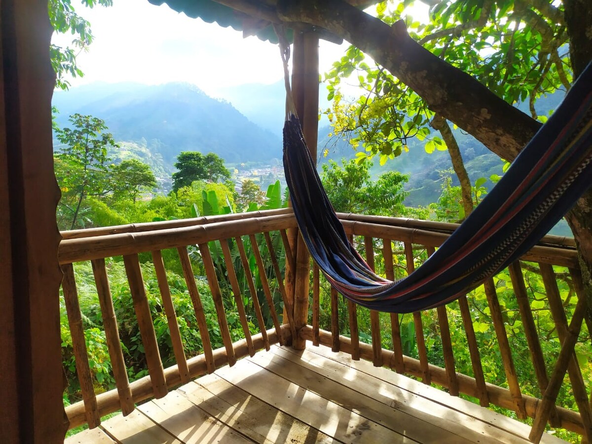 A shaded wooden deck features a vibrant hammock, gently swaying between wooden posts. Lush greenery fills the surrounding area, and distant mountains create a serene backdrop, contributing to a peaceful outdoor setting.