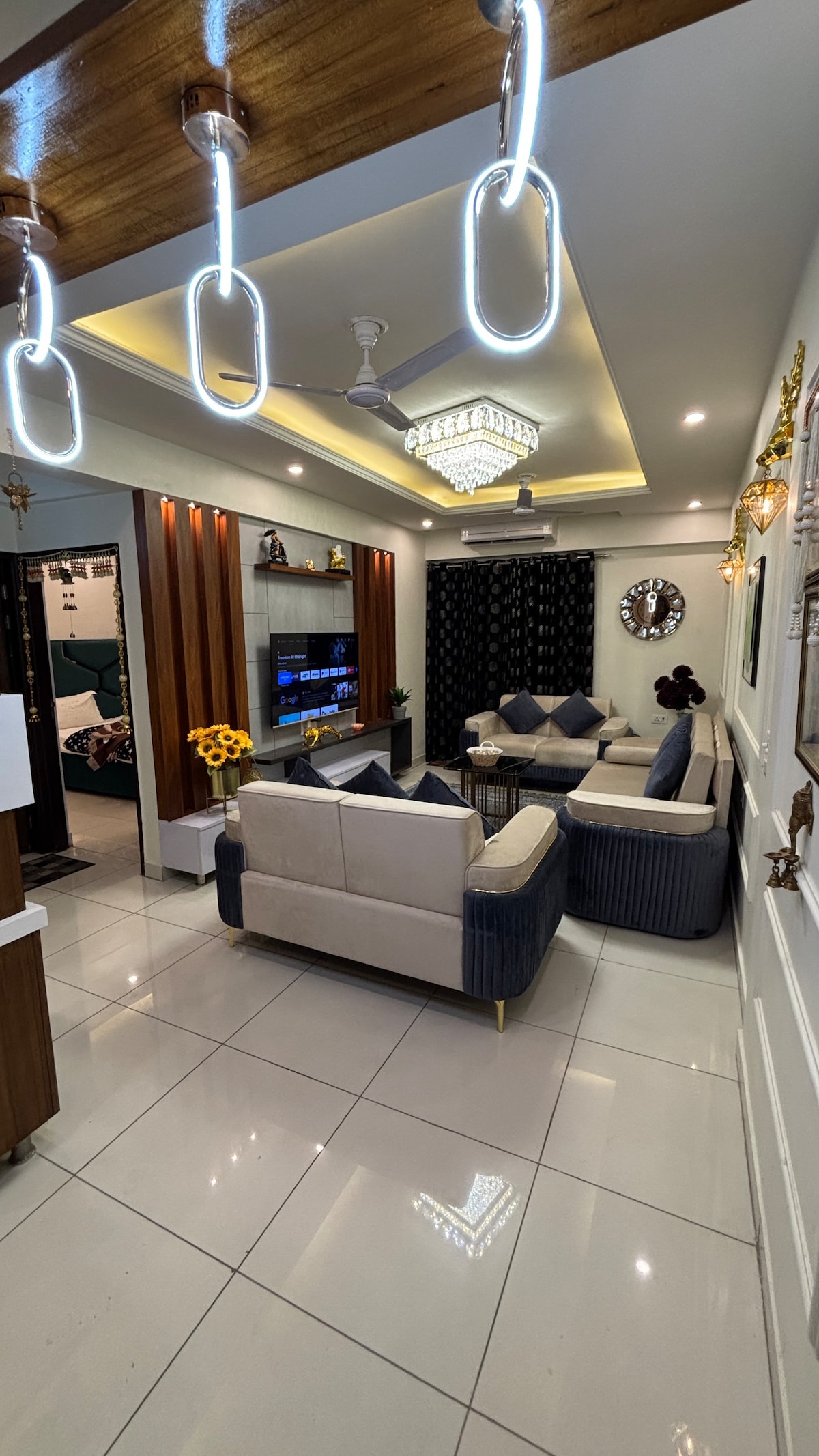 A modern living area is highlighted by a crystal chandelier and unique lighting fixtures. A comfortable seating arrangement of light-colored sofas with dark blue accent cushions surrounds a coffee table. A television is mounted on a feature wall, complementing the sleek tile flooring.