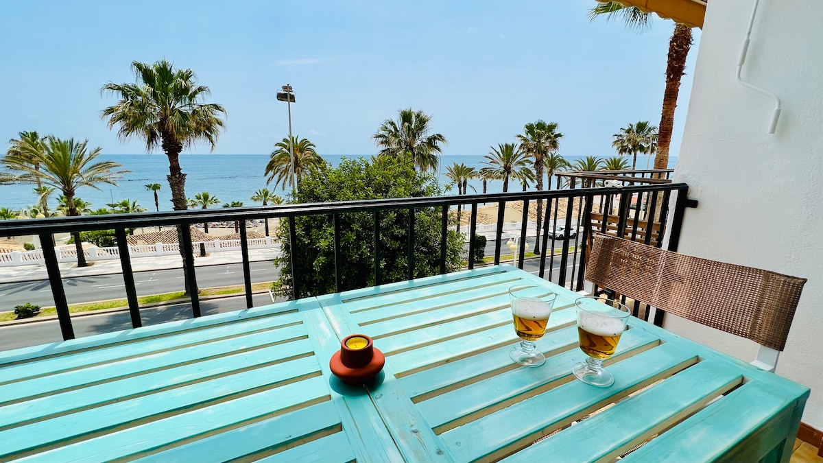 A balcony table, painted in a soft turquoise, is set with two glasses of beer and a small candle. The view showcases palm trees framing a sandy beach and the ocean, with a road visible in the foreground.
