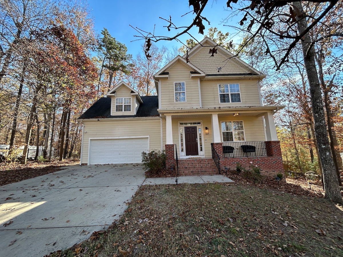 A two-story house is set amongst a backdrop of trees with autumn leaves. The exterior features light-colored siding, a black roof, and red brick accents. A driveway leads to a two-car garage, and a welcoming front porch extends toward the entrance.