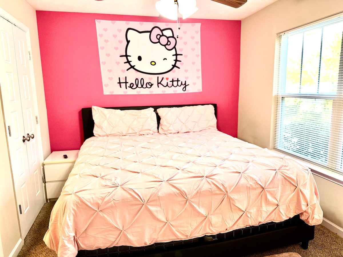 A sleeping area features a large, plush bed with a pink quilted coverlet. A cheerful Hello Kitty wall tapestry adorns a bright pink accent wall. A nightstand is positioned beside the bed, and a window allows natural light to fill the room.