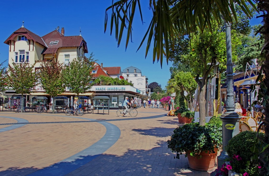 A vibrant street scene is depicted, featuring a wide pathway lined with potted plants and trees. Several people can be seen walking or biking, while outdoor seating areas are arranged near cafés and shops. The clear blue sky enhances the lively atmosphere.