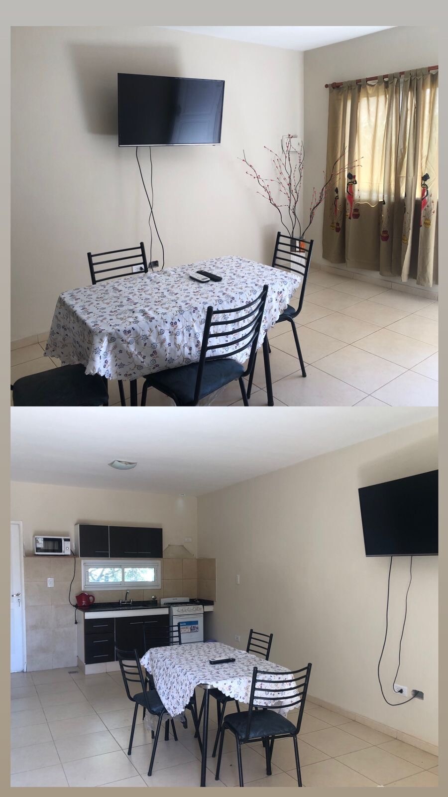 A dining area is showcased with a table dressed in a floral tablecloth and surrounded by black chairs. A television is mounted on the wall, and a kitchen space with dark cabinetry is visible in the background, complemented by light-colored walls.
