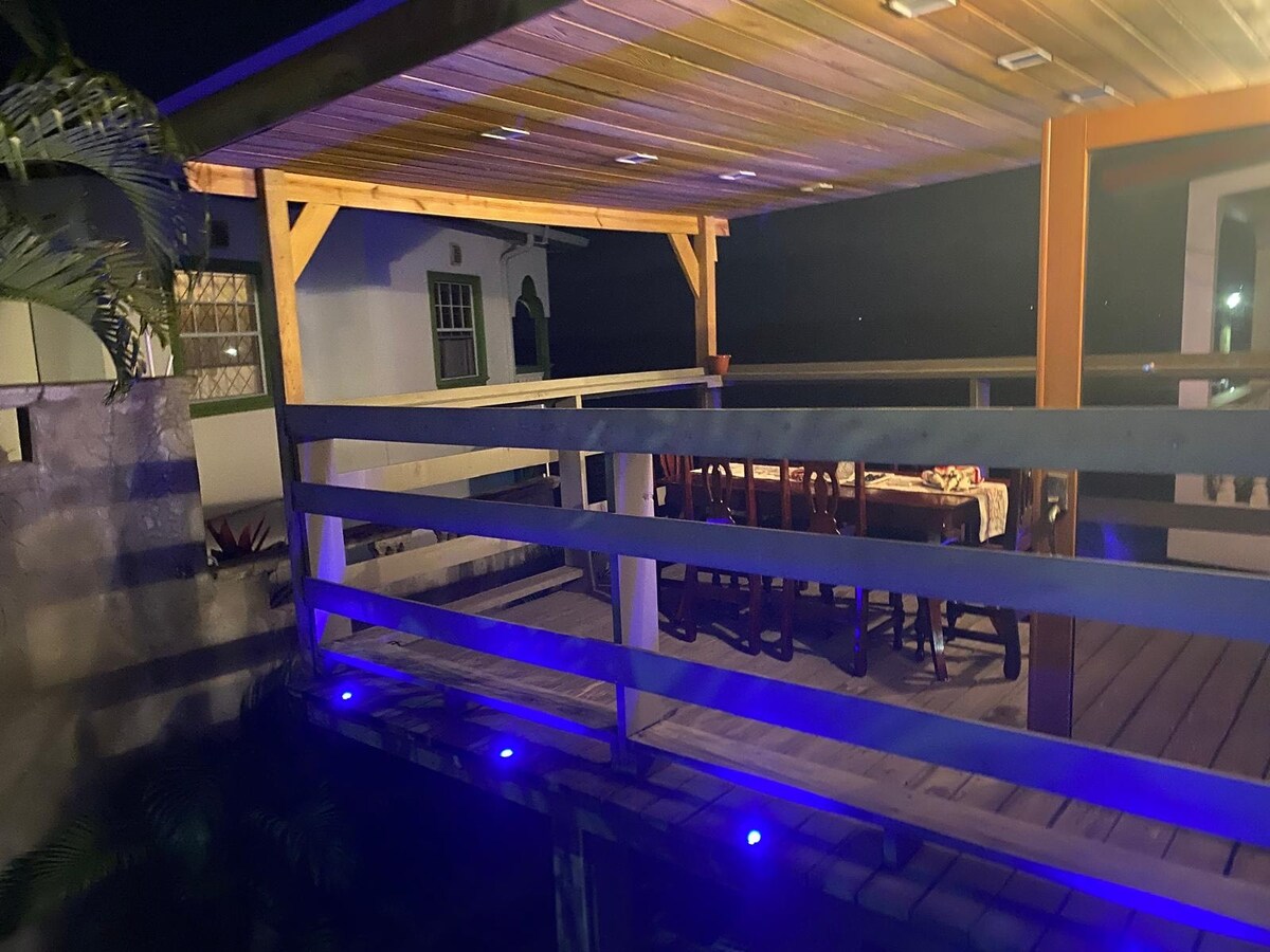 A spacious outdoor dining area is situated beneath a wooden canopy, illuminated by subtle blue lighting. A long table with several chairs invites gatherings. The surrounding area features lush greenery, enhancing the serene atmosphere during evening hours.