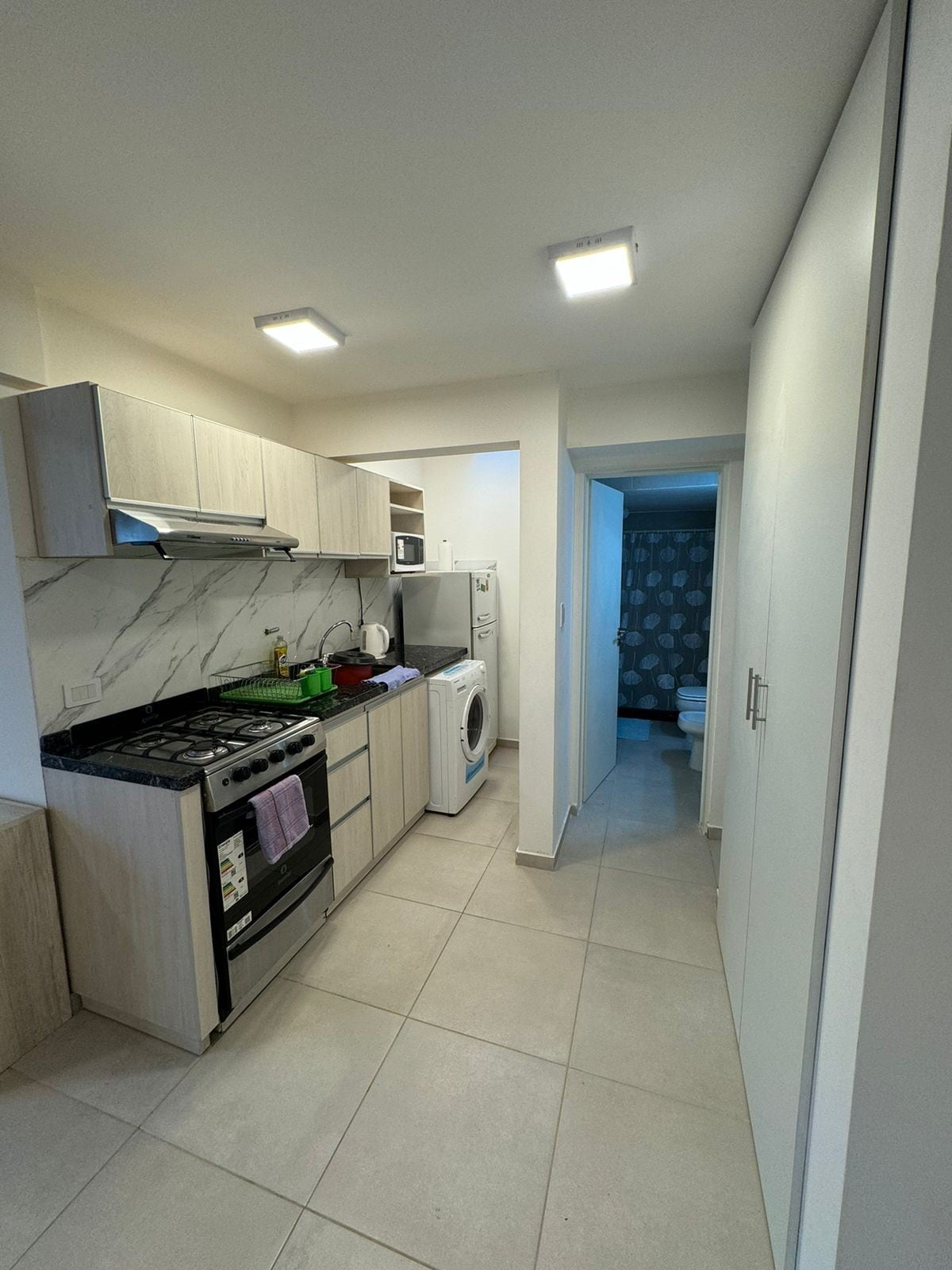 The kitchen area features modern cabinetry with a marble backsplash, equipped with a stove, microwave, and refrigerator. A washer is positioned beside the appliances. Bright overhead lighting illuminates the tiled floor, while a doorway leads to the adjacent bathroom.