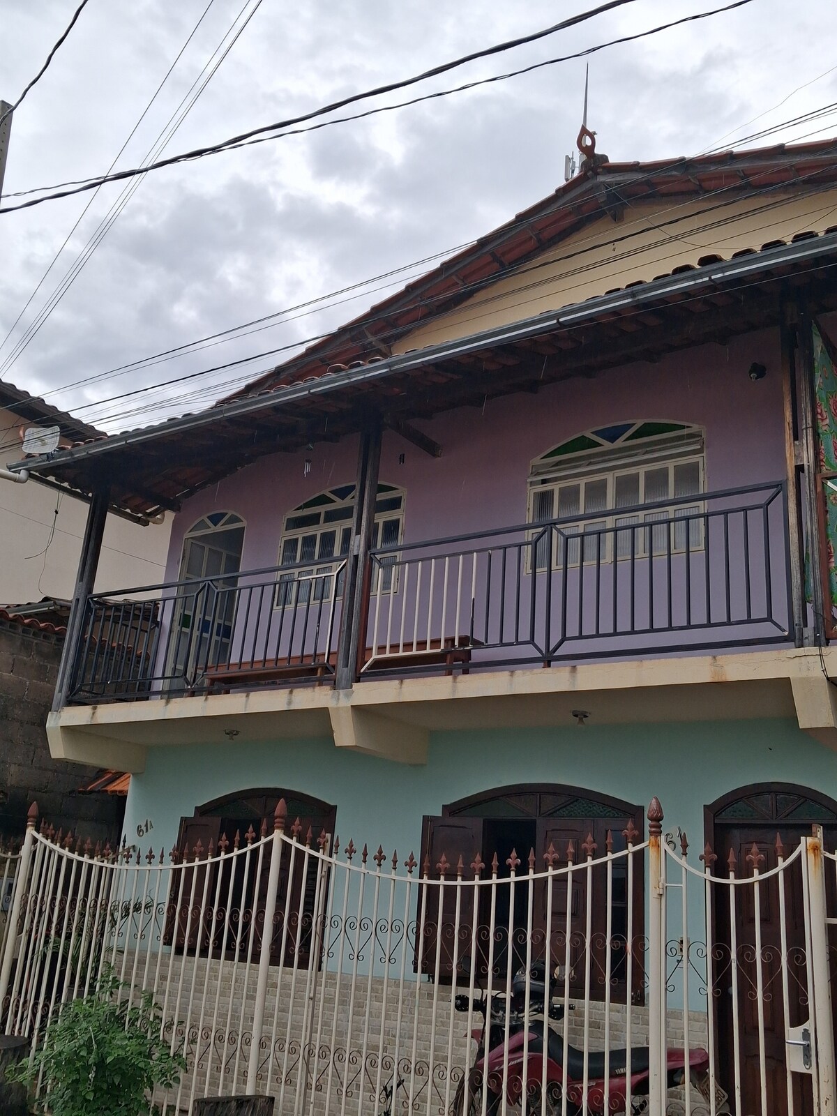 The exterior of a two-story building is visible, featuring a light purple facade and a welcoming balcony with black railings. The front entrance has multiple doors under a roofed area, while a decorative iron fence encloses the property, providing a sense of privacy and security.