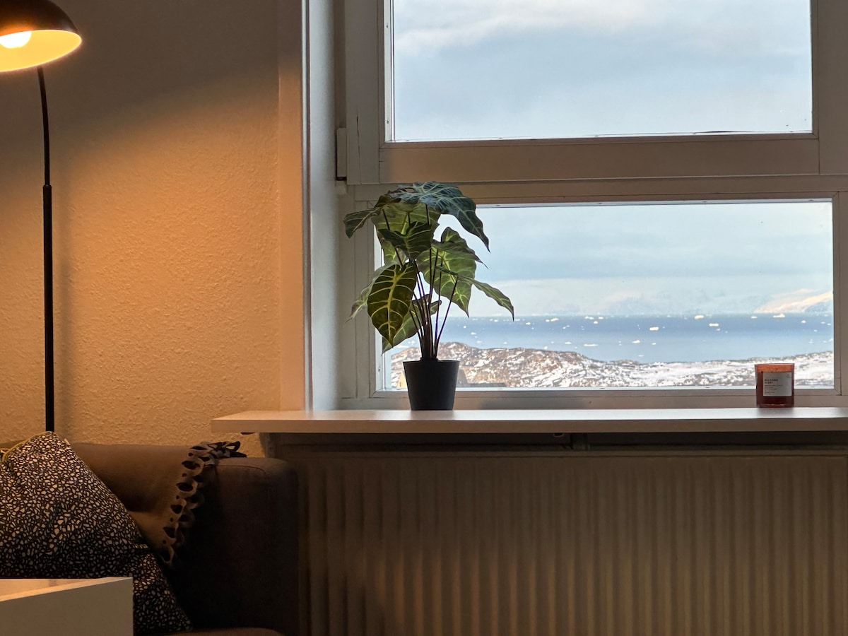 A window offers a view of the stunning Diskobugten and distant icebergs. A potted plant sits on the ledge, complementing a cozy sofa nearby. A lamp provides soft lighting, creating a peaceful ambiance for relaxation.