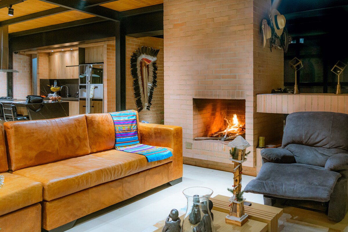 The living space features a spacious leather sofa with colorful textiles, complemented by a comfortable armchair. A well-defined fireplace is visible, providing a warm focal point. The exposed brick walls and wooden beams enhance a cozy ambiance, while glimpses of the kitchen area are seen in the background.