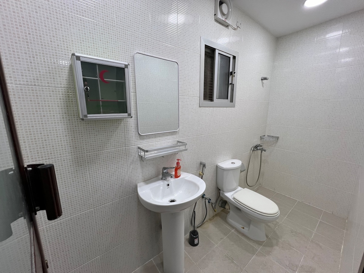 The bathroom features a clean design with light-colored tiles covering the walls and floor. A white sink with a mirrored cabinet is positioned next to a modern toilet. Natural light enters through a small window, and a shower area is located in the corner.