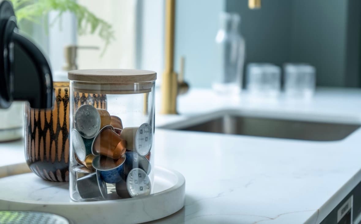 A clear glass jar containing various coffee capsules is placed on a smooth, light-colored kitchen countertop. A minimalist plate holds the jar, while a stylish black faucet and a sink are visible in the background. Soft natural light adds a warm touch to the scene.