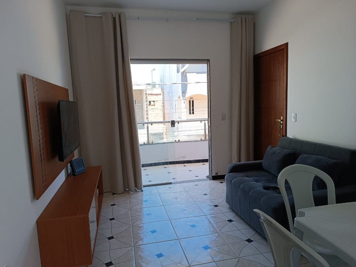 A bright living area is presented with a gray sofa bed facing a wooden TV stand. Large windows allow natural light to fill the space, with light curtains partially drawn. A small white table and chairs are positioned nearby, enhancing the room's functionality.