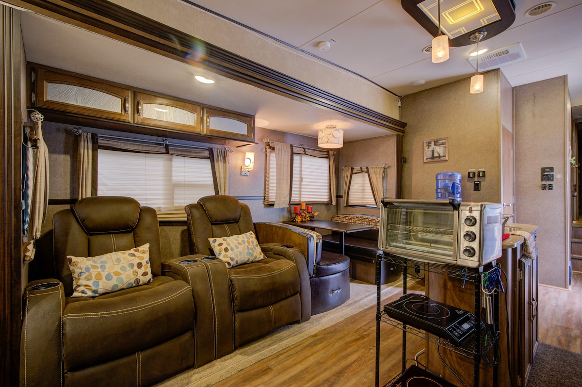 The living area features two comfortable reclining chairs, each adorned with decorative pillows. A compact kitchen area is visible, equipped with a microwave and a water cooler. Natural light enters through large windows covered with light-filtering shades, creating a cozy atmosphere.