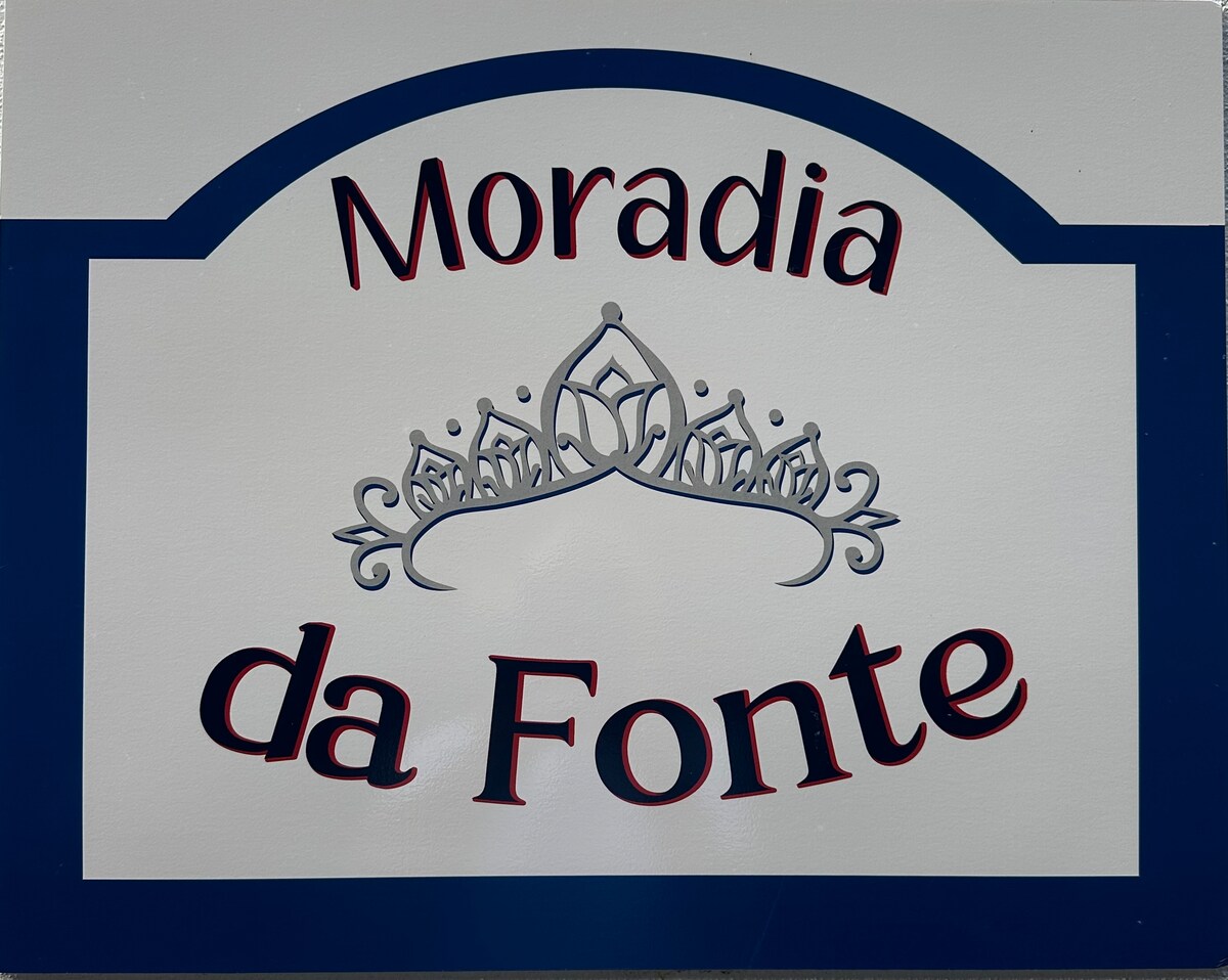 A sign displays the name 'Moradia da Fonte,' featuring an ornate crown design above the text. The background is a light grey with a blue border, offering a clear and inviting identification of the property.