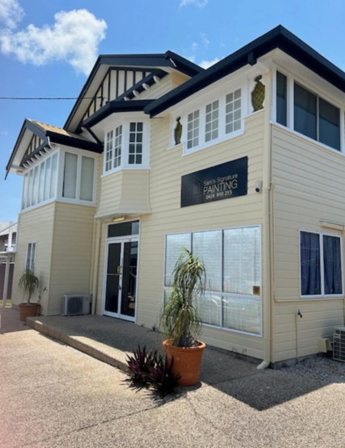 A two-story structure features a light yellow exterior with decorative architectural elements. Large windows and a glass door create a bright entrance, while potted plants add a touch of greenery to the front. The signage indicates a local painting business.