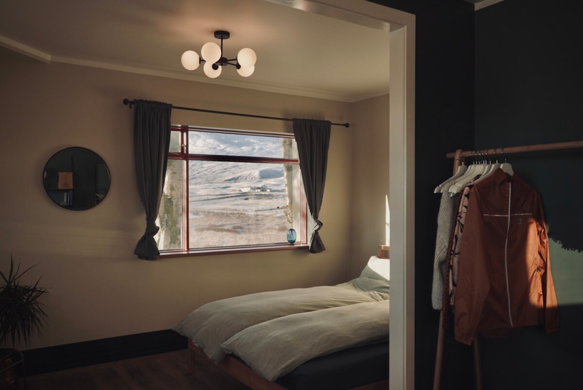 A spacious bedroom features a large window framed by soft curtains, inviting natural light and offering scenic views of the landscape outside. A comfortable bed is adorned with neutral bedding, while a clothing rack holds a few garments, creating an uncluttered and serene atmosphere.