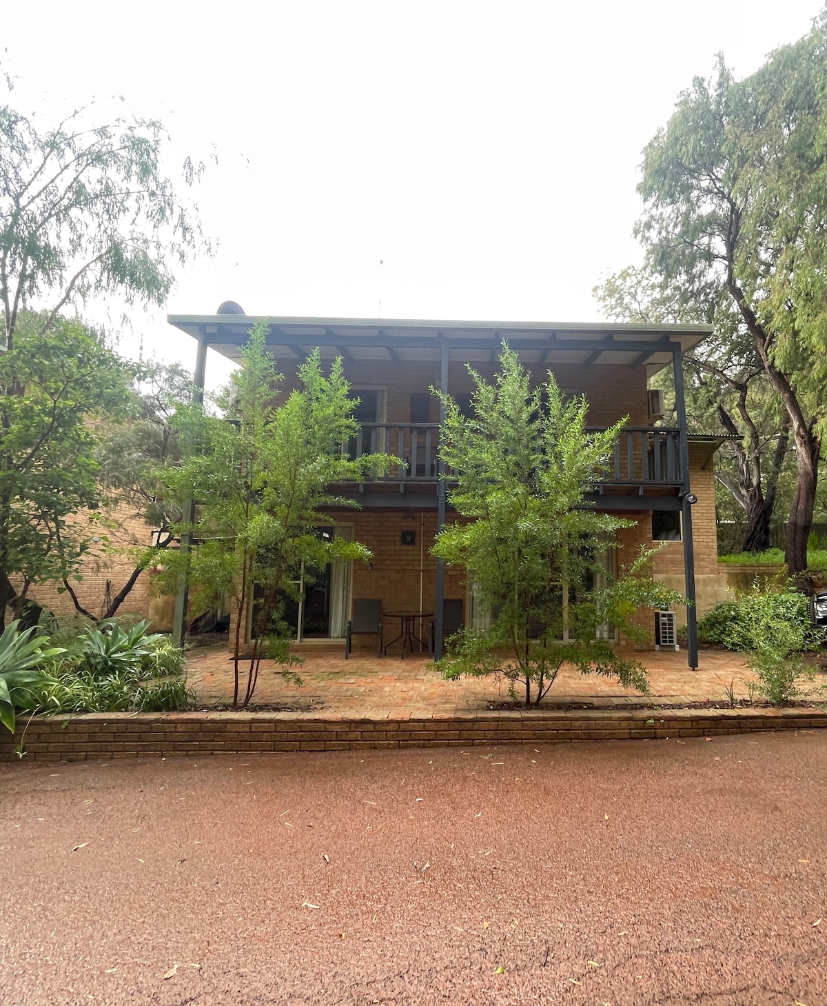 The chalet's exterior presents a two-story structure surrounded by greenery. A covered balcony is visible on the upper level, providing access to tree-top views. The entrance features a pathway lined with natural plants, creating a serene approach to the retreat.
