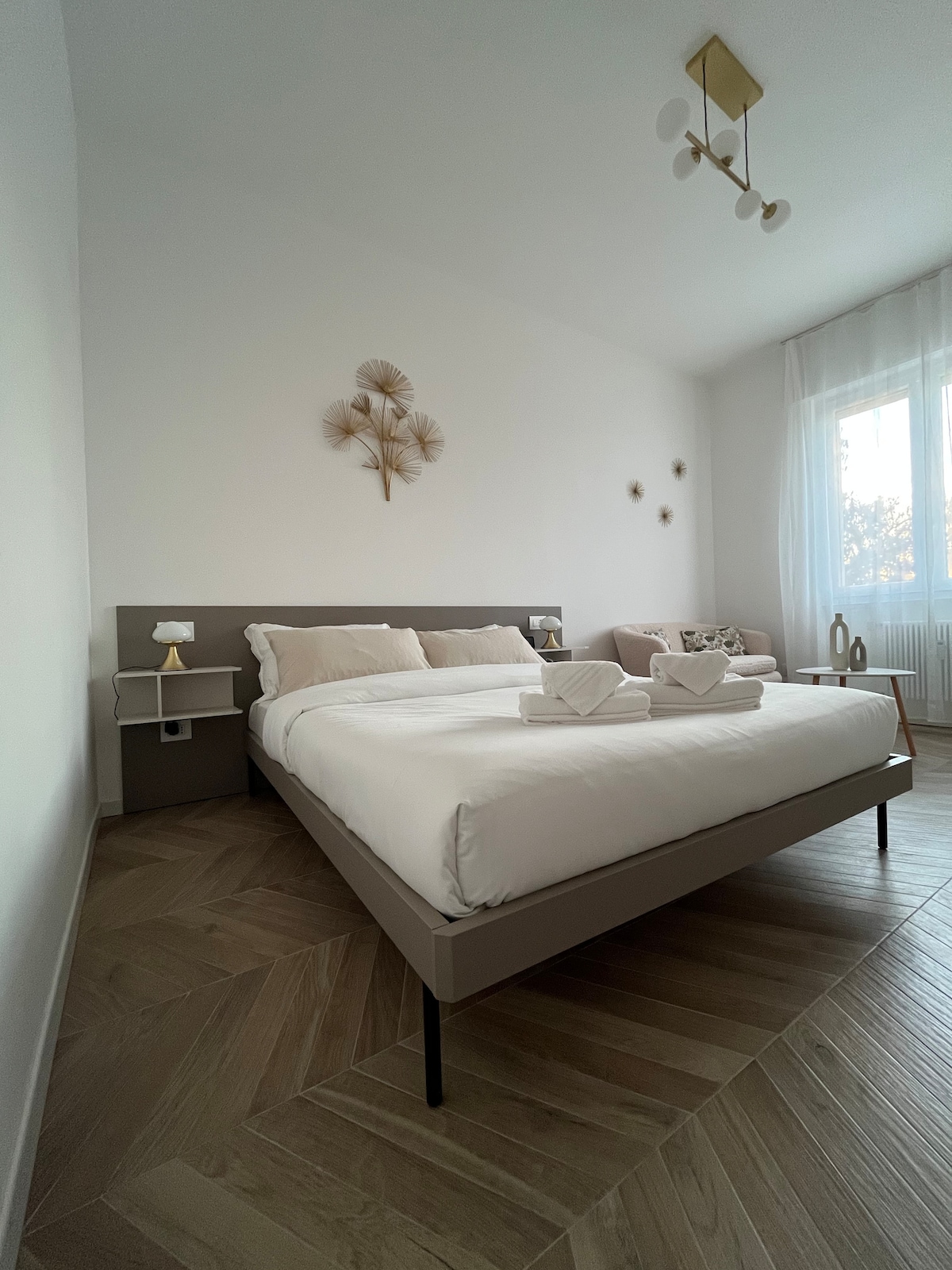 A spacious bedroom features a large bed with light bedding and decorative pillows. Natural light filters through sheer curtains, enhancing the room's calming ambiance. A small side table and a cozy seating area with a sofa complement the elegant decor, creating a welcoming environment.