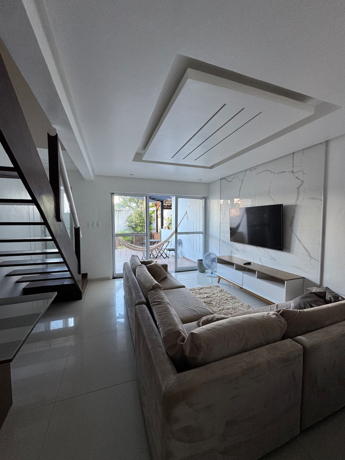 A modern living area features a comfortable sectional sofa and a wall-mounted television. Large glass doors lead to a patio, allowing natural light to fill the space. A staircase, enhanced by a sleek design, is visible on one side of the room.